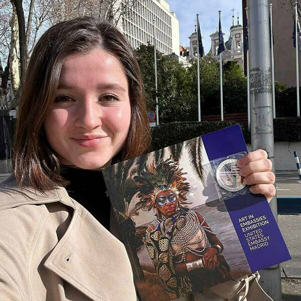 Raleigh Kuipers after her meeting with Ambassador Julissa Reynoso, holding the Ambassador's Challenge Coin and a booklet about the art collection in the embassy. The U.S. Embassyin Madrid is visible in the background.